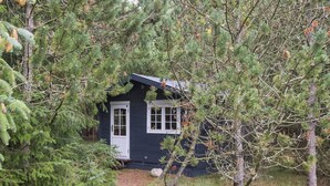 A small cabin with a door and windows, surrounded by dense trees and a gravel path. - 5 Star Holiday Home in Blavand (Blavand)