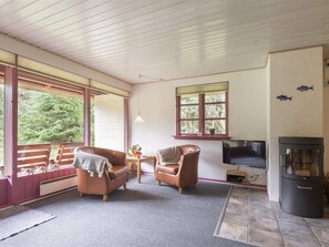 Ein gemütliches Wohnzimmer mit Kamin, zwei Ledersesseln, einem Fernseher und einem großen Fenster mit Blick ins Grüne.