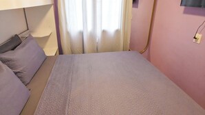 Ein Schlafzimmer mit einem Bett, einem Kopfteil, einem Fenster mit Vorhängen und einem an der Wand befestigten Fernseher.