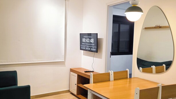Ein Zimmer mit Holztisch und -stühlen, einem kleinen Schrank, einem Fernseher, der die Uhrzeit anzeigt, und einem Spiegel an der Wand.