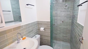 Ein Badezimmer mit Duschkabine aus Glas, einem weißen Waschbecken mit Seifenschale, einer Toilette und einem Handtuchhalter.