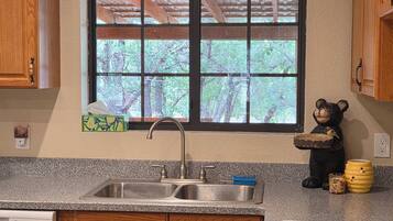 A kitchen with wooden cabinets, a granite countertop, a sink, a window with a view of trees, and a bear figurine on the counter.