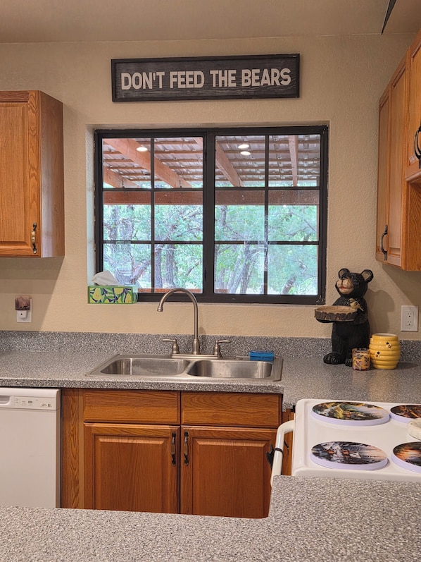 A kitchen with wooden cabinets, a granite countertop, a sink, a window with a view of trees, and a bear figurine on the counter.