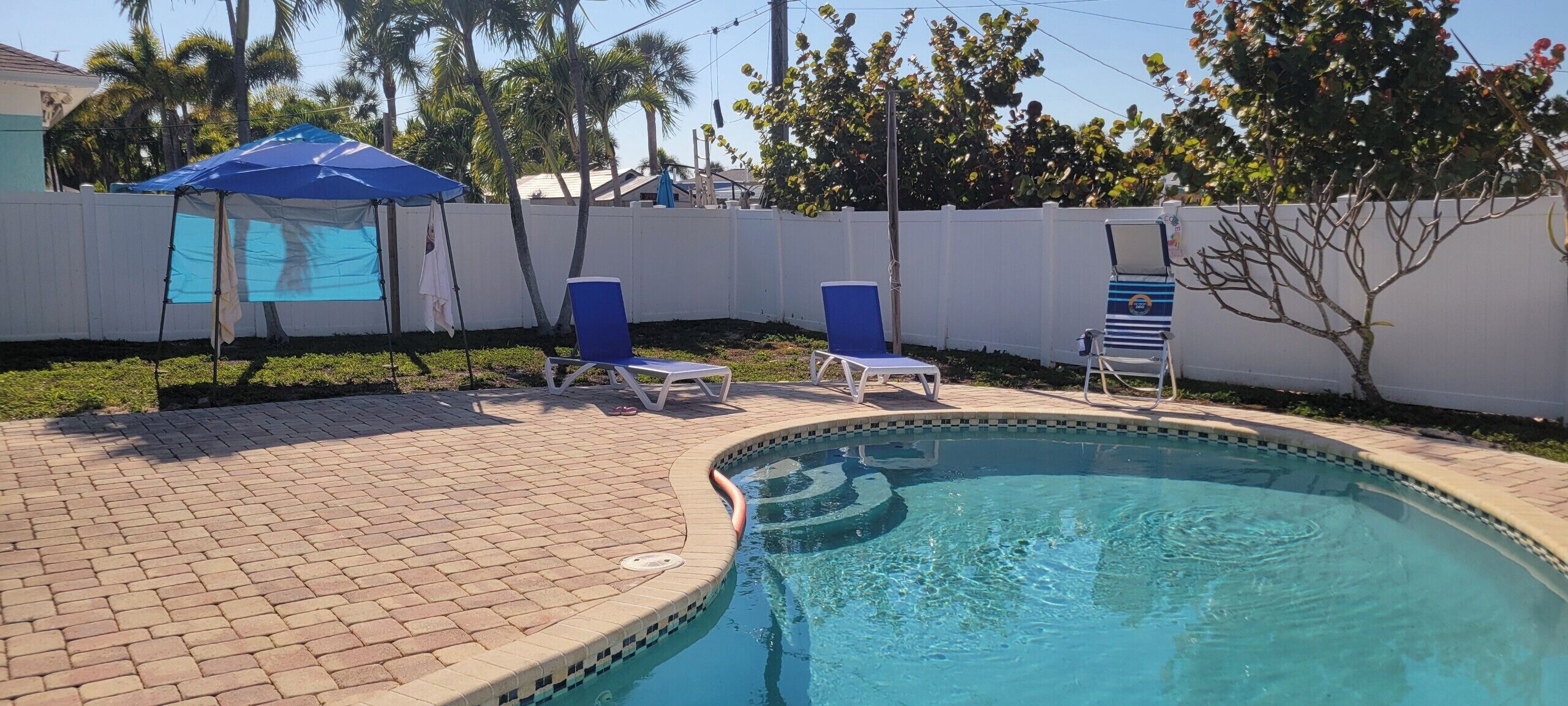 Une piscine dont le bord est recouvert de carreaux, entourée d’une terrasse en briques. On y trouve des chaises longues et un parasol bleu.