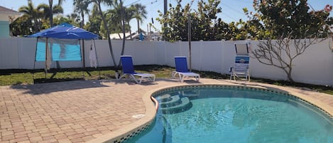 Une piscine dont le bord est recouvert de carreaux, entourée d’une terrasse en briques. On y trouve des chaises longues et un parasol bleu.