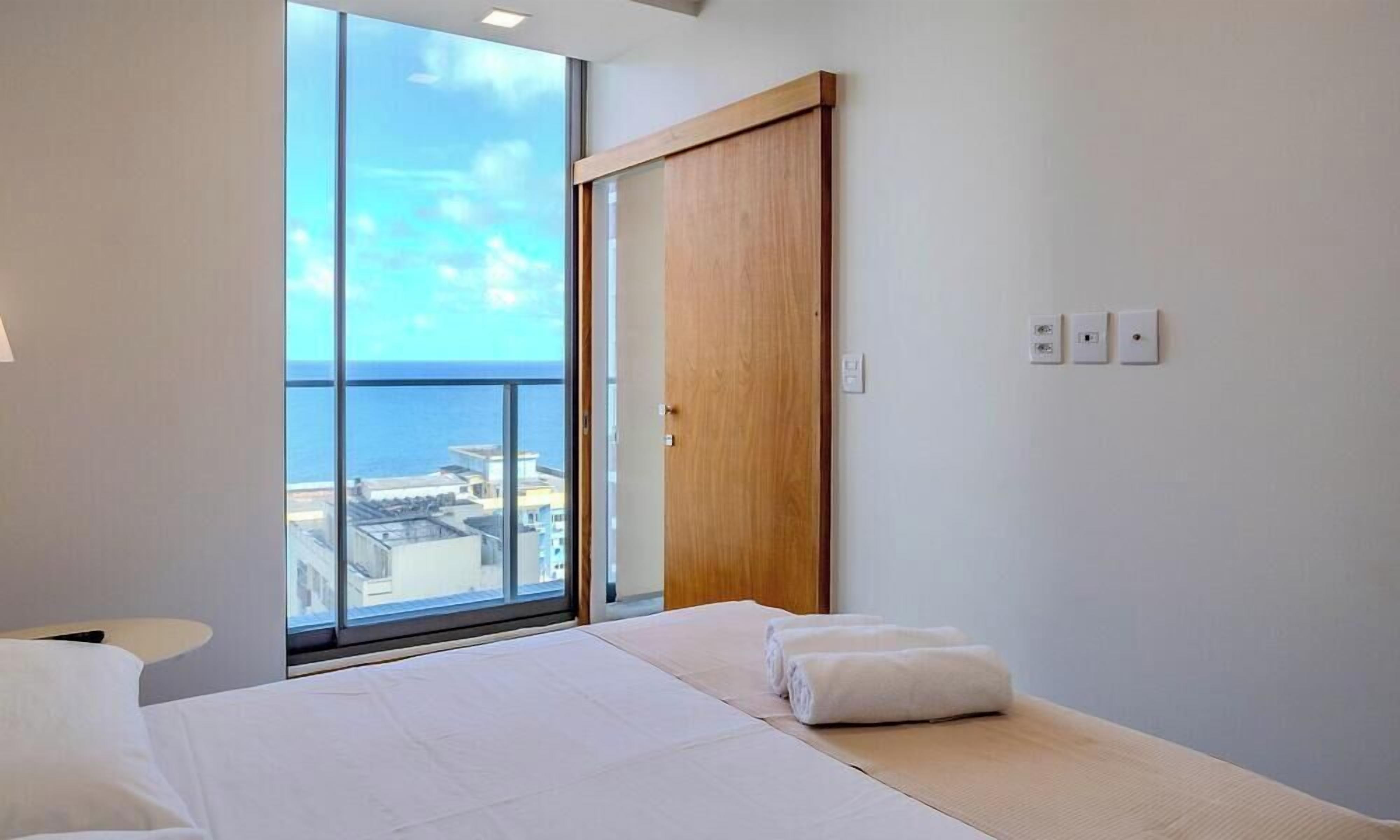 A hotel room with a large window, a bed, a wooden door, and a view of the sea.