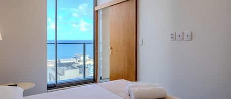 A hotel room with a large window, a bed, a wooden door, and a view of the sea.