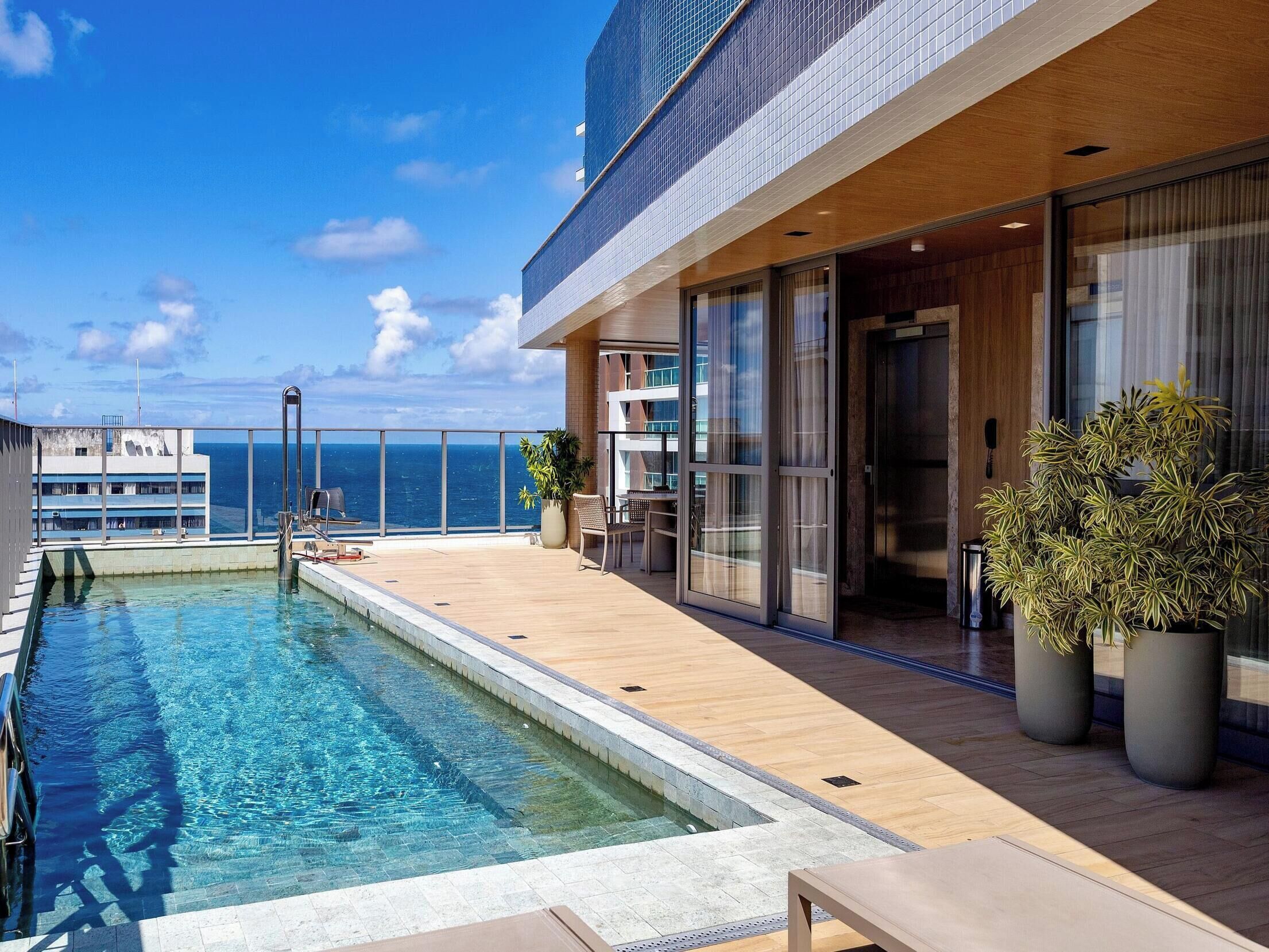 A poolside area with a clear view of the ocean, a wooden deck, and a building with large windows.