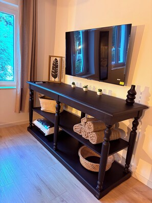 A dark wooden console table with a flat-screen TV mounted above it, a framed leaf decoration, and a small potted plant. - Le Mocopek Vacation Rental (sleeps 5) (Bolbec)
