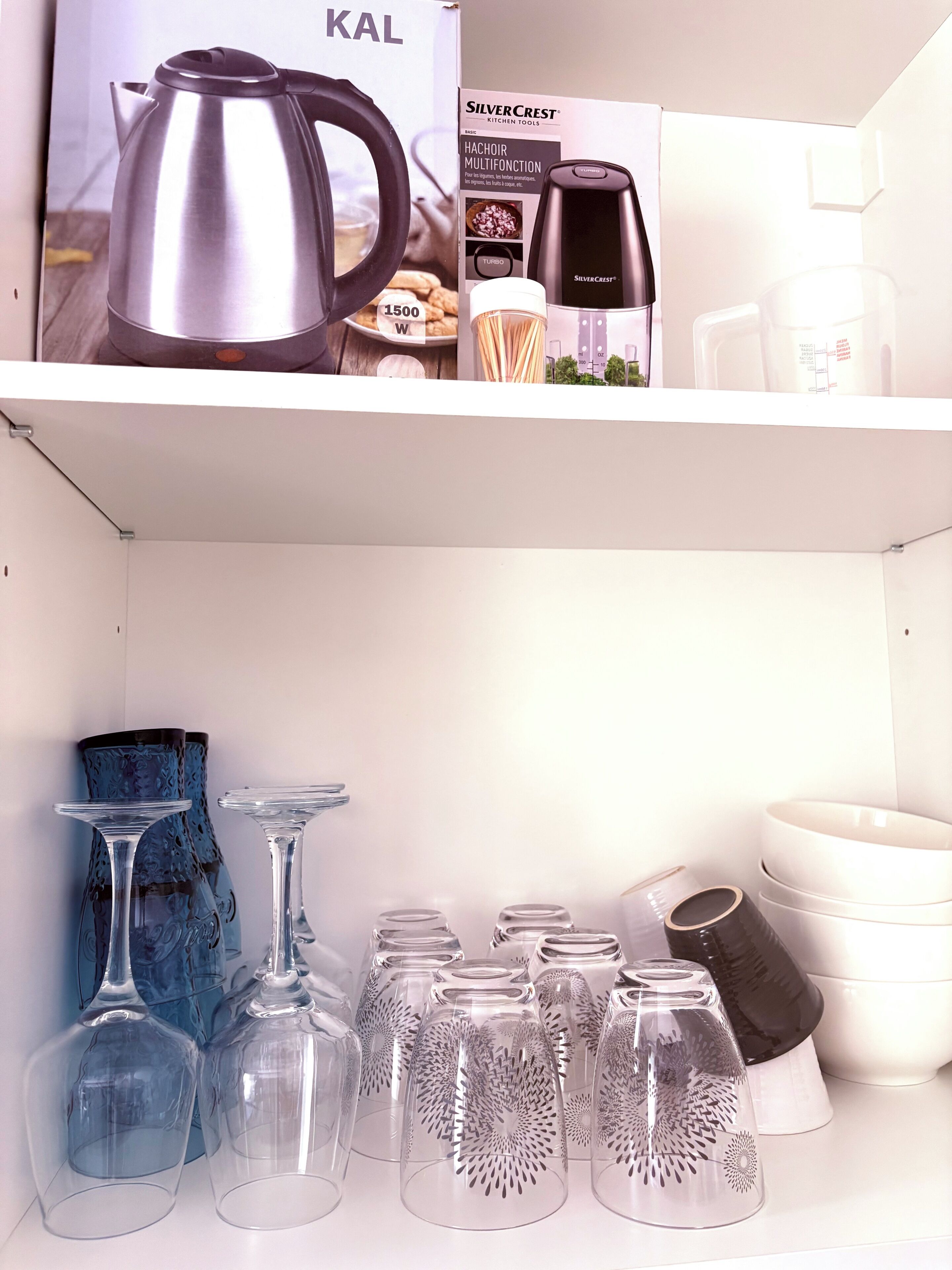 Una estantería de cocina con un hervidor, una cafetera y una caja de utensilios. Abajo, hay copas de vino y un florero.