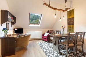 Ein gemütliches Zimmer mit Esstisch, Stühlen, einem Fernseher, einem Fenster mit Blick auf Bäume und einer dekorativen Pendelleuchte.