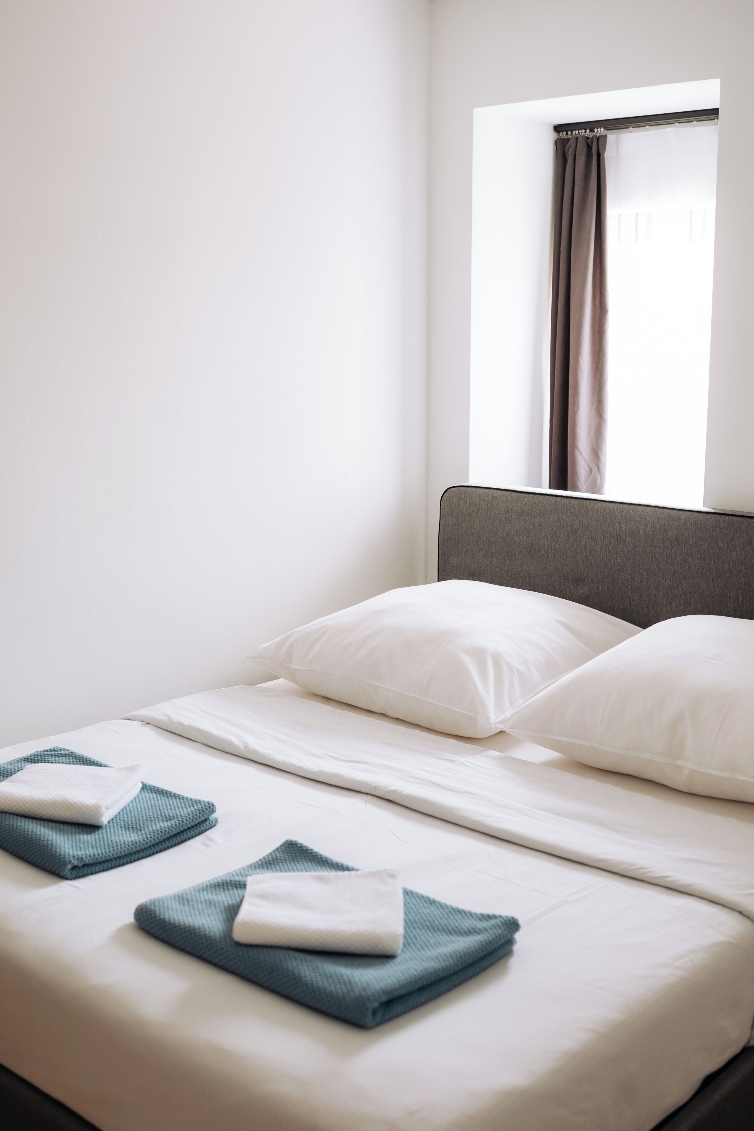 Un lit bien fait, avec du linge de lit blanc et deux serviettes pliées, une tête de lit grise et une fenêtre ornée d’un rideau marron.
