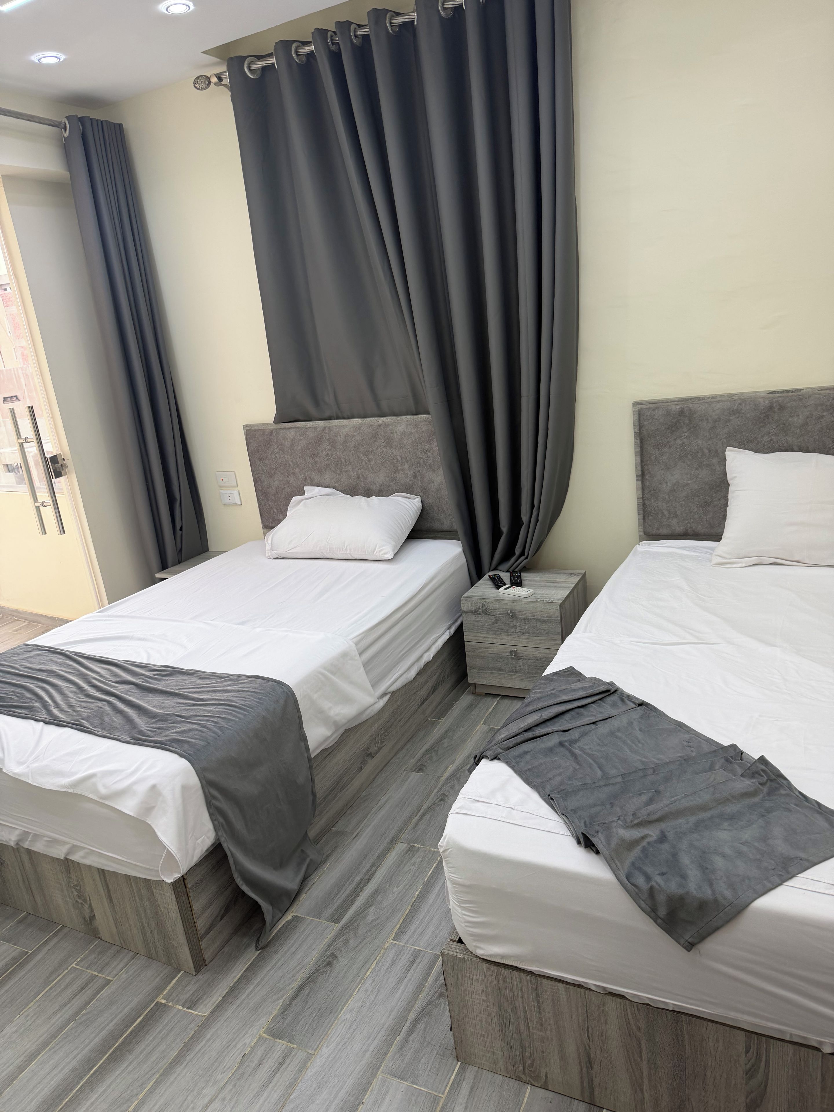 Una habitación de hotel con dos camas, cortinas grises, una mesita de noche con lámpara y suelo de madera.