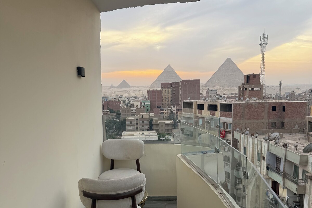 A balcony with a chair, a glass railing, and a view of pyramids in the distance.