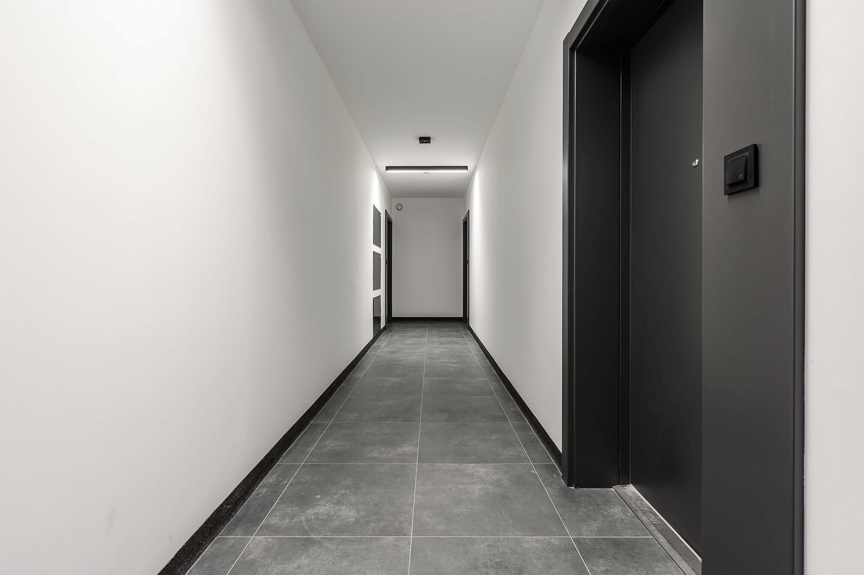 A long, narrow hallway with a dark door on the right and a light-colored wall on the left.