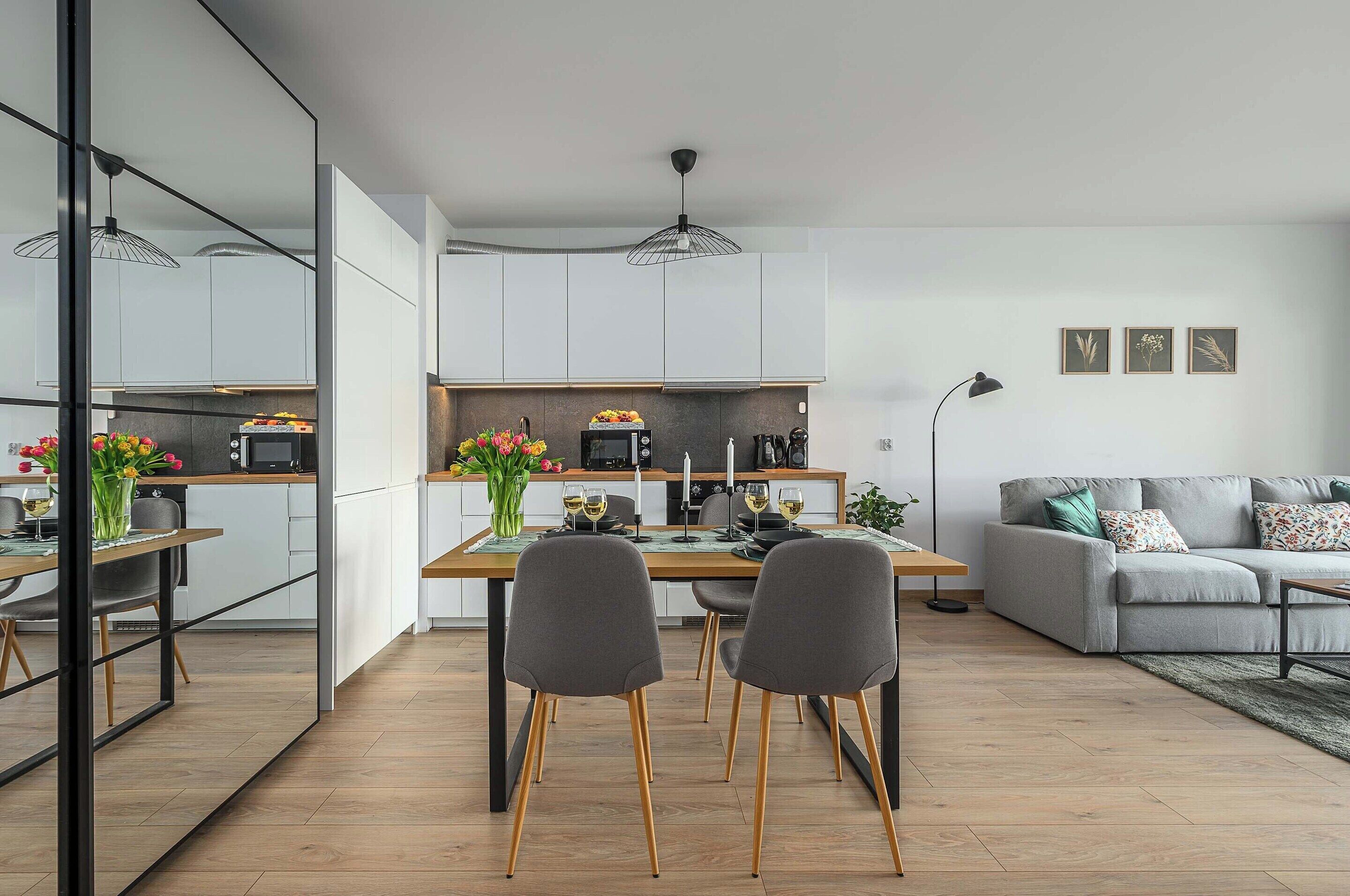 A modern kitchen with white cabinets, a dining area with two chairs, and a grey sofa in a living room.