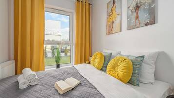 A bedroom with a bed, a window with curtains, a radiator, a plant, and artwork on the wall.