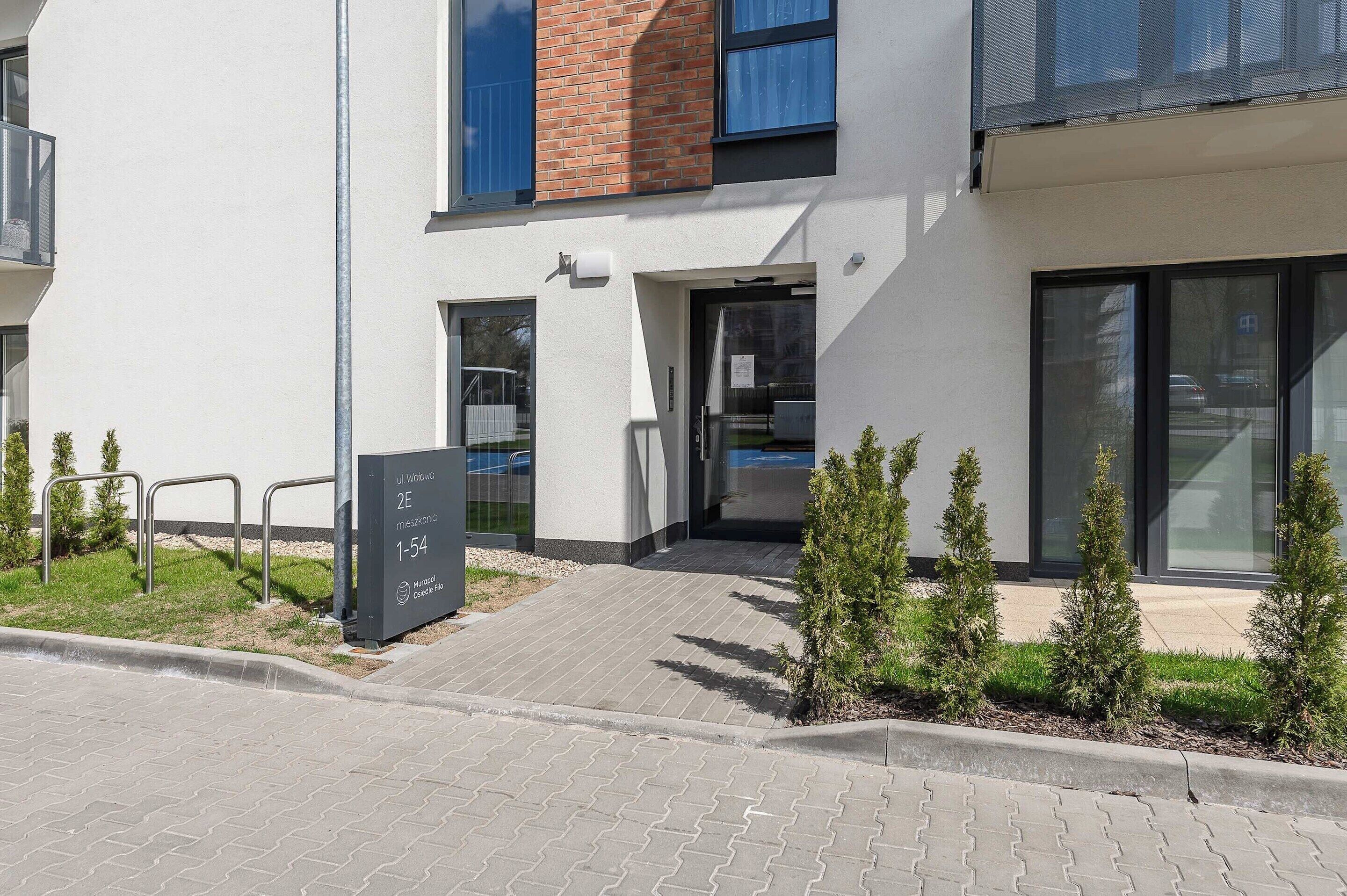 Een modern gebouw met een bakstenen gevel, grote ramen en een verharde entreezone met kleine bomen.