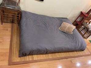 Ein Bett mit grauem Matratze und einem braunen Kopfkissen, Holzboden und ein Sessel mit Sitzkissen.