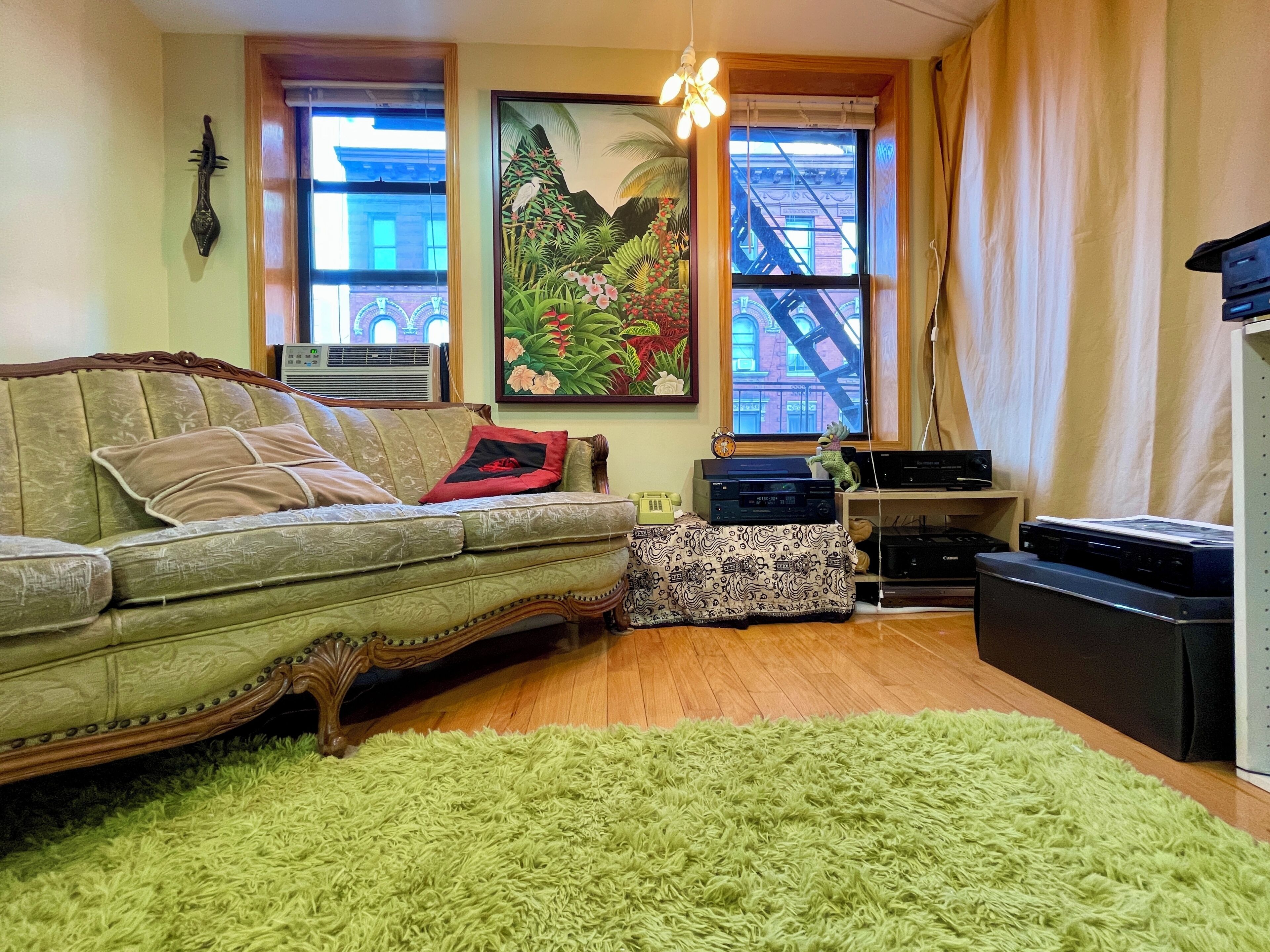 A living room with a vintage sofa, a green rug, a painting, and a view of a building through a window.