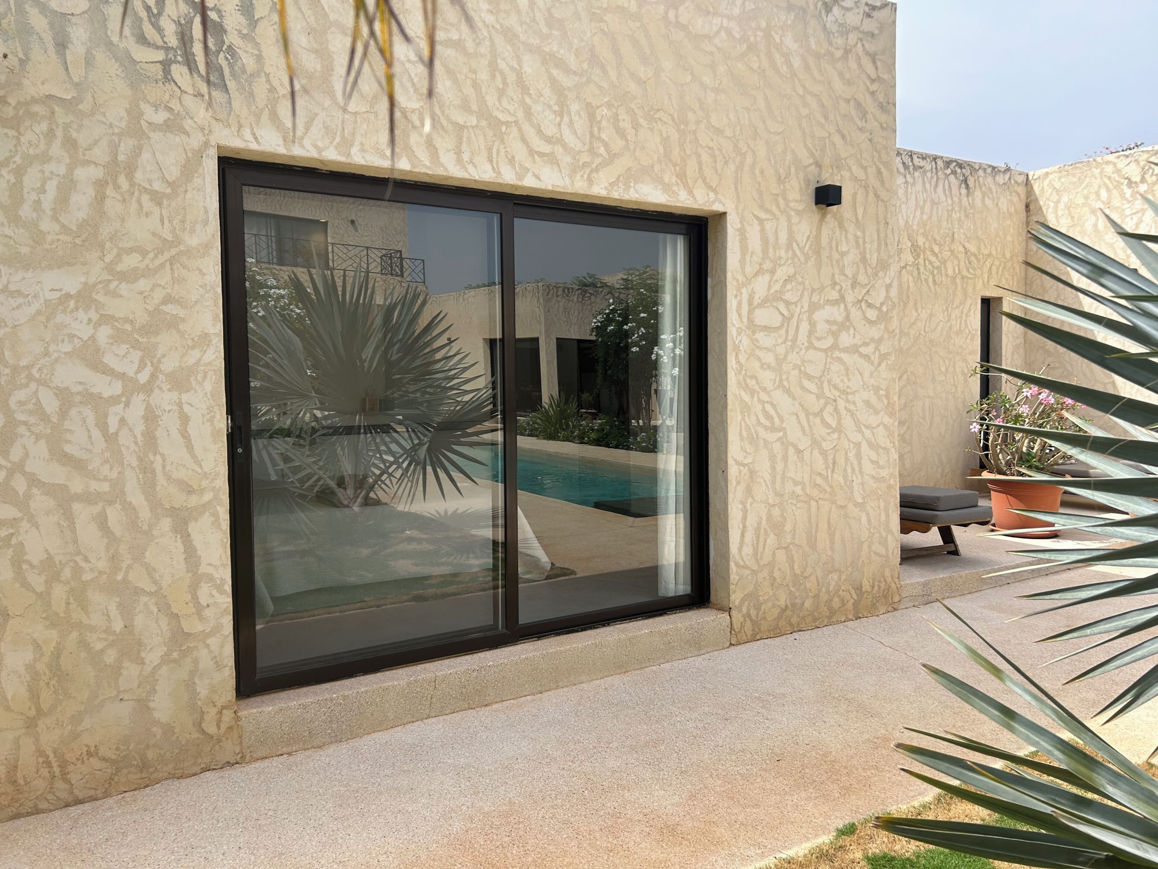 Un edificio moderno con un amplio ventanal corredera, piscina y patio con una silla y plantas en macetas.