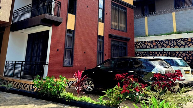 A multi-story residential building with balconies, a car parked in front, and a paved driveway.