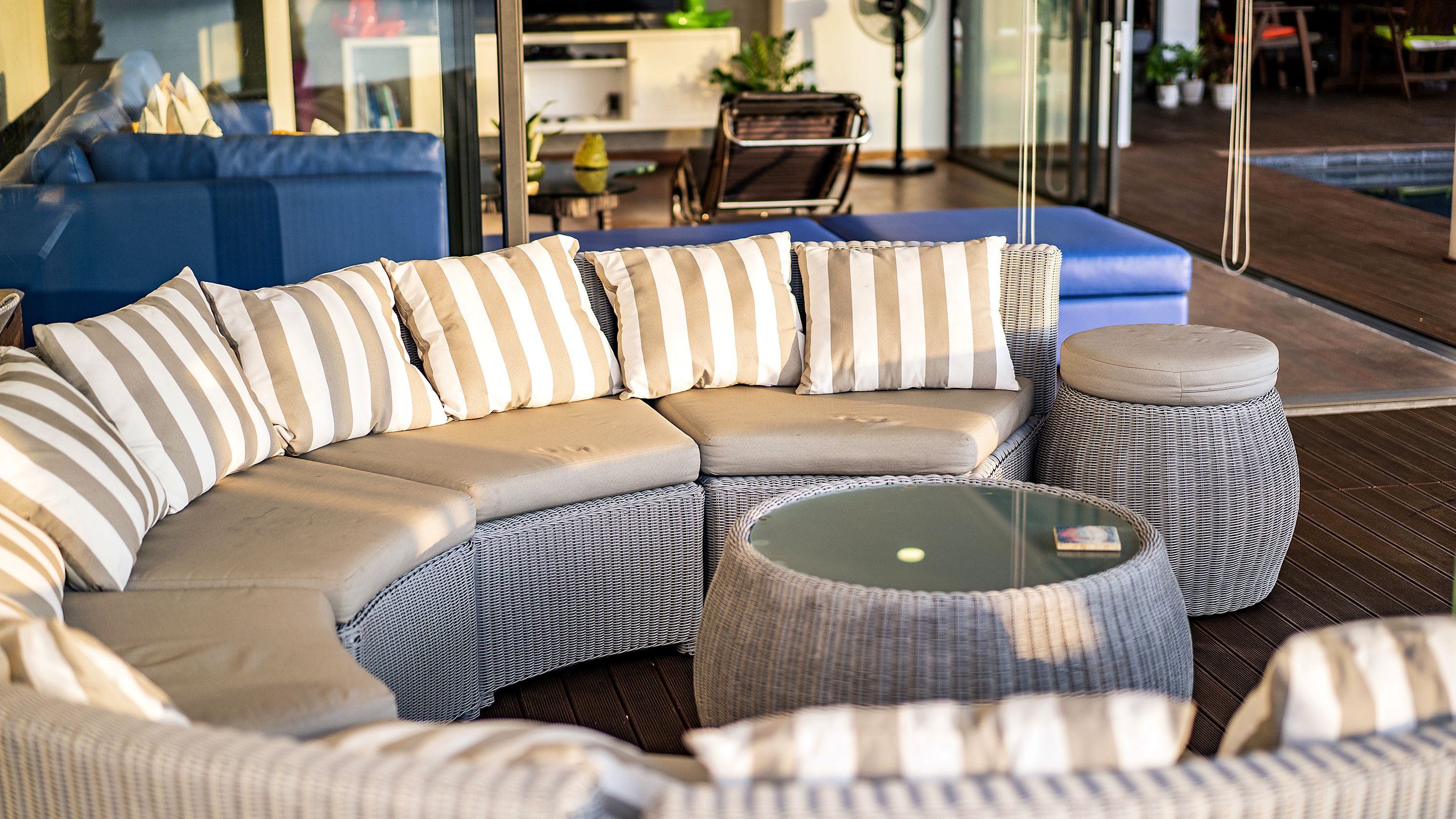 A sectional sofa with striped cushions, a round wicker ottoman, and a glass-top coffee table in a modern outdoor setting.