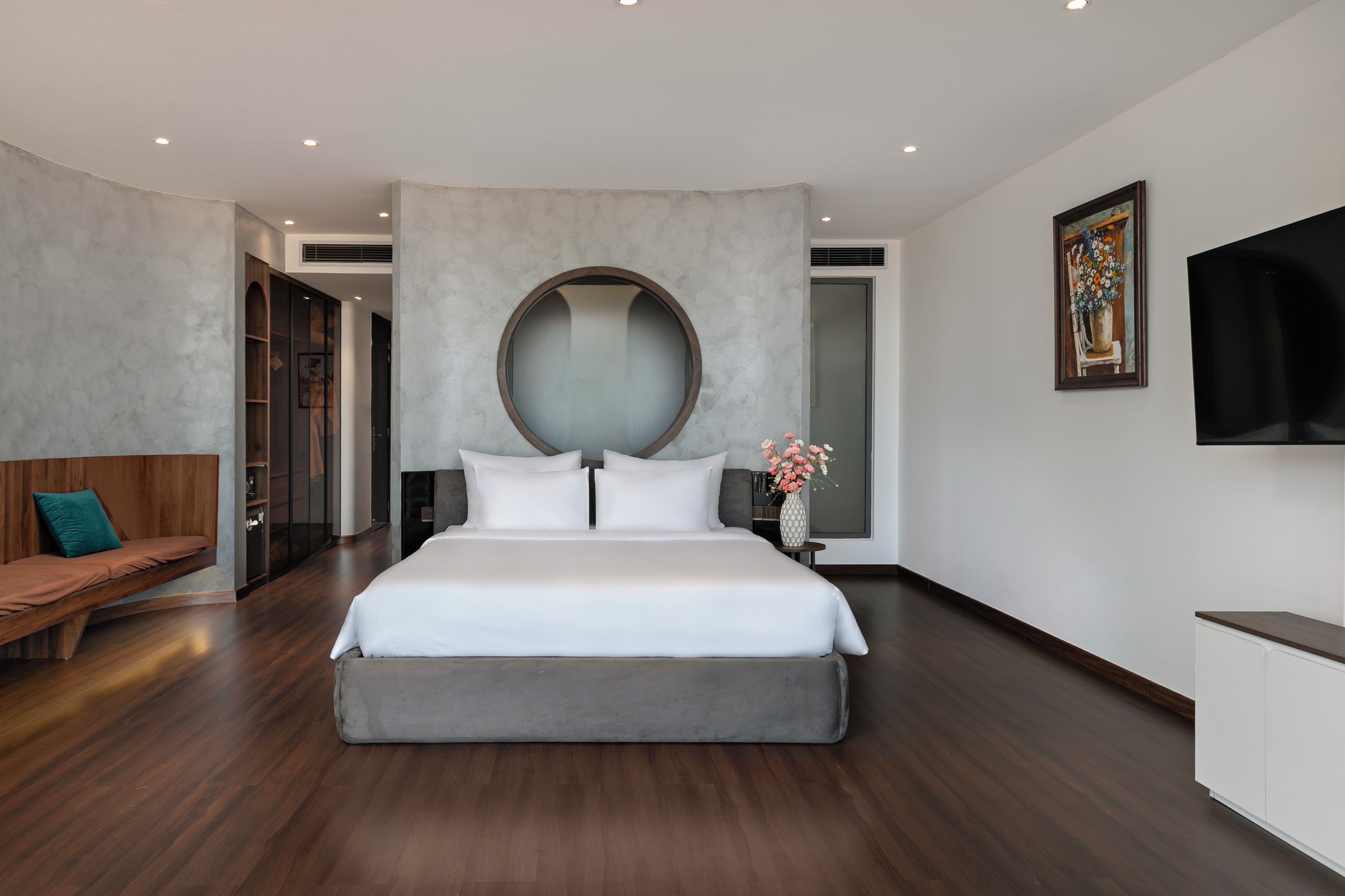 Un dormitorio moderno con una cama grande, un espejo redondo, un banco de madera y un televisor de pantalla plana montado en la pared.