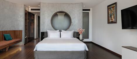 A modern bedroom with a large bed, a round mirror, a wooden bench, and a flat-screen TV mounted on the wall.