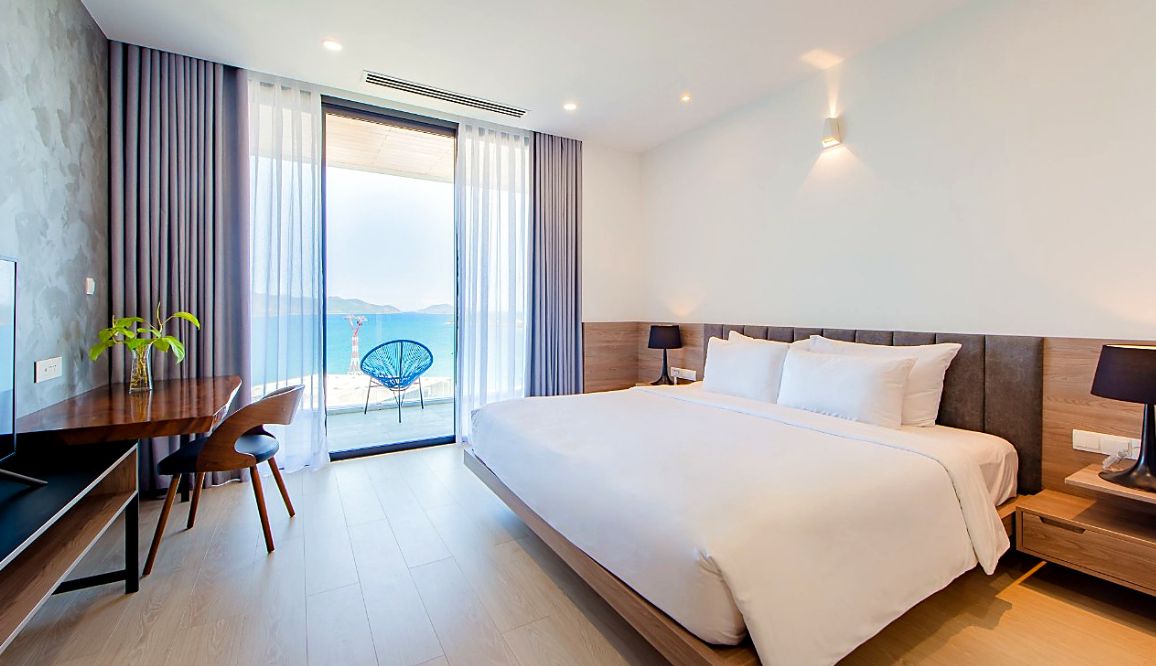 A modern hotel room with a large bed, a desk, and a chair. There is a view of the sea through the window.