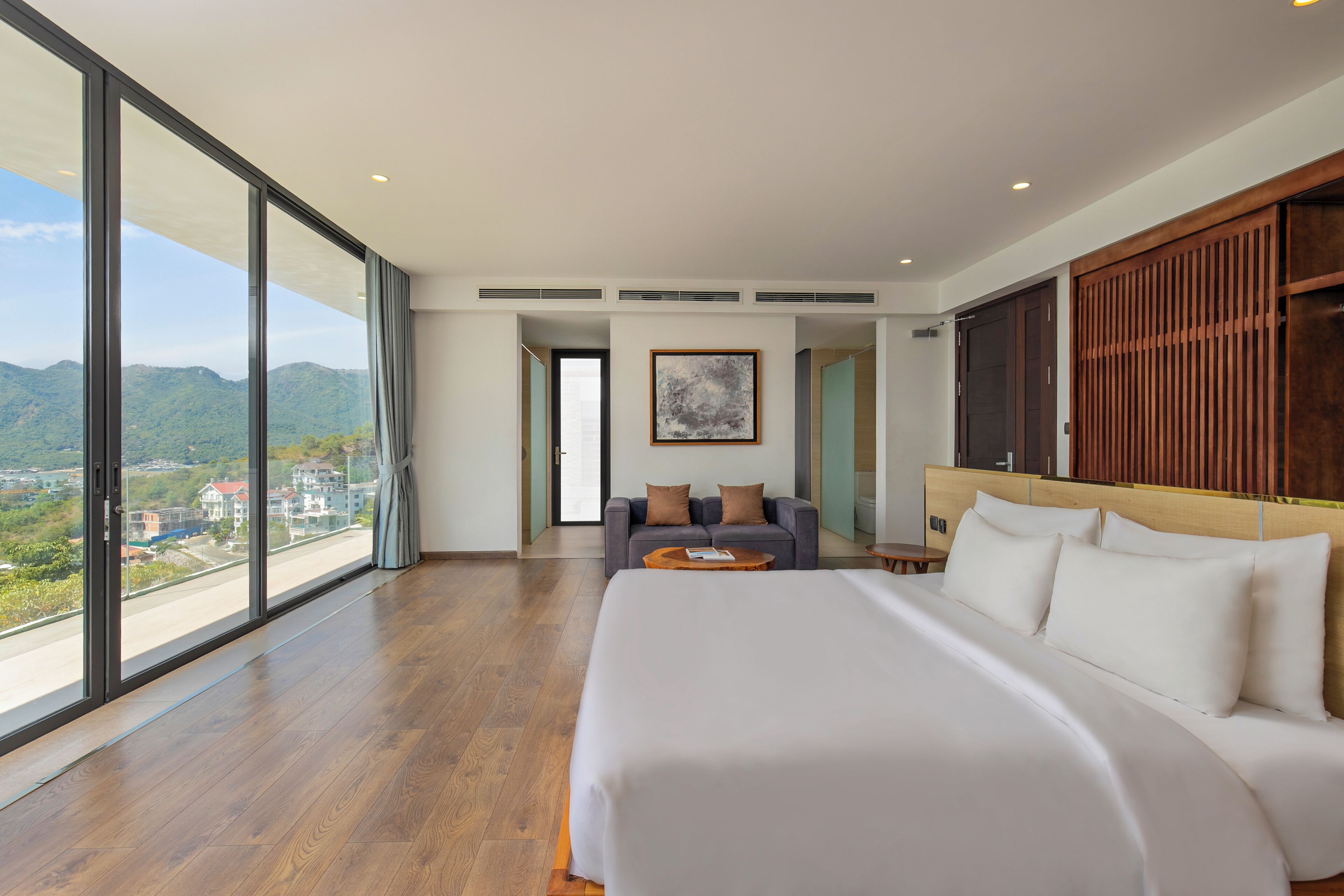 A modern hotel room with a large bed, a seating area with a sofa, and a view of the mountains through floor-to-ceiling windows.