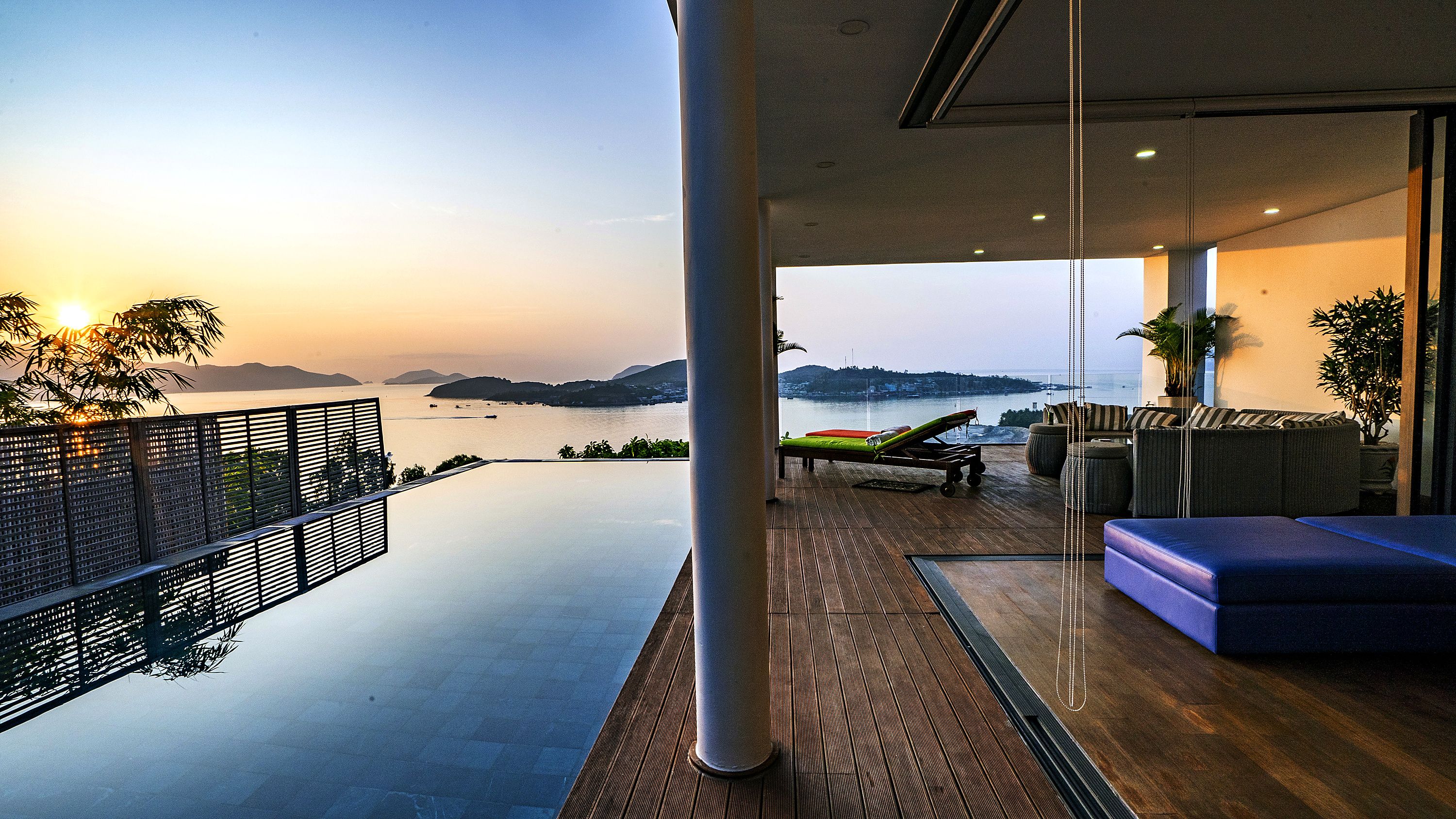 A modern balcony with a view of the sea and distant hills, a lounge chair, and a sofa.