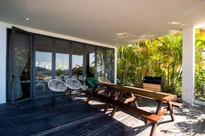 Un patio con mesa y sillas de madera, rodeado por puertas y ventanas de vidrio, con vista a la vegetación.