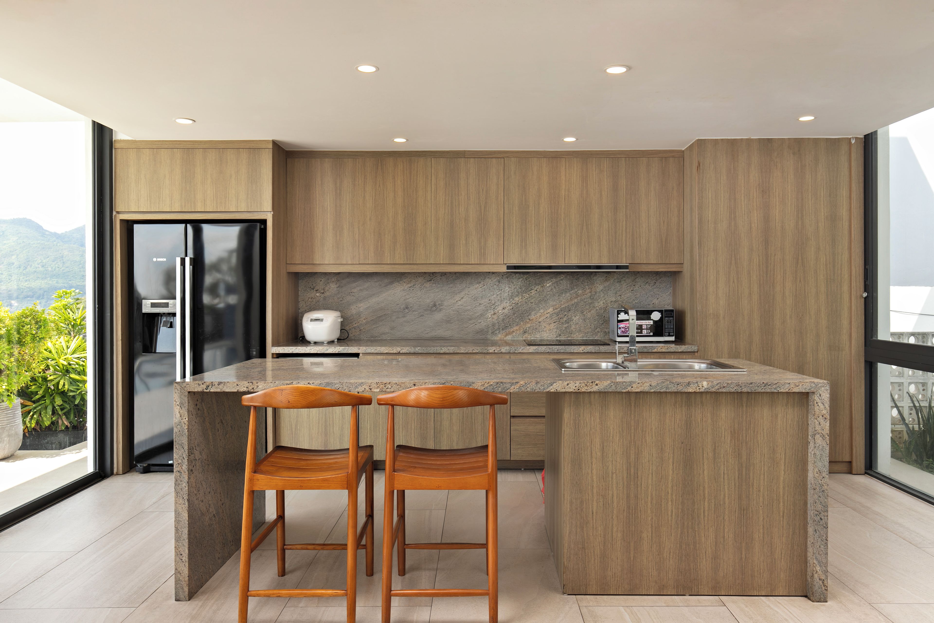 A modern kitchen with wooden cabinetry, a granite countertop, and stainless steel appliances. There are two wooden bar stools at the island.
