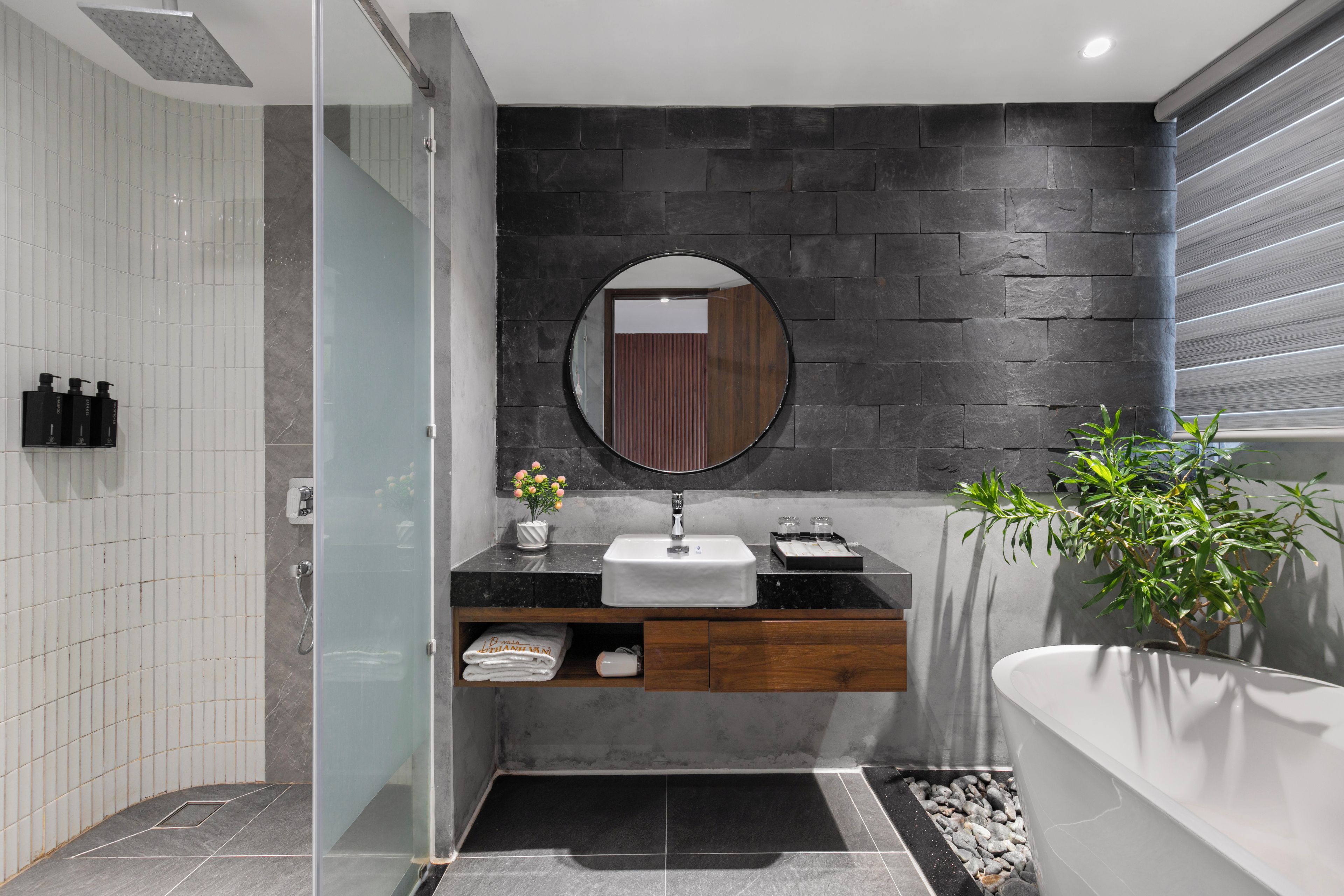 A modern bathroom with a glass shower enclosure, a round mirror, a sink with a wooden vanity, and a potted plant.