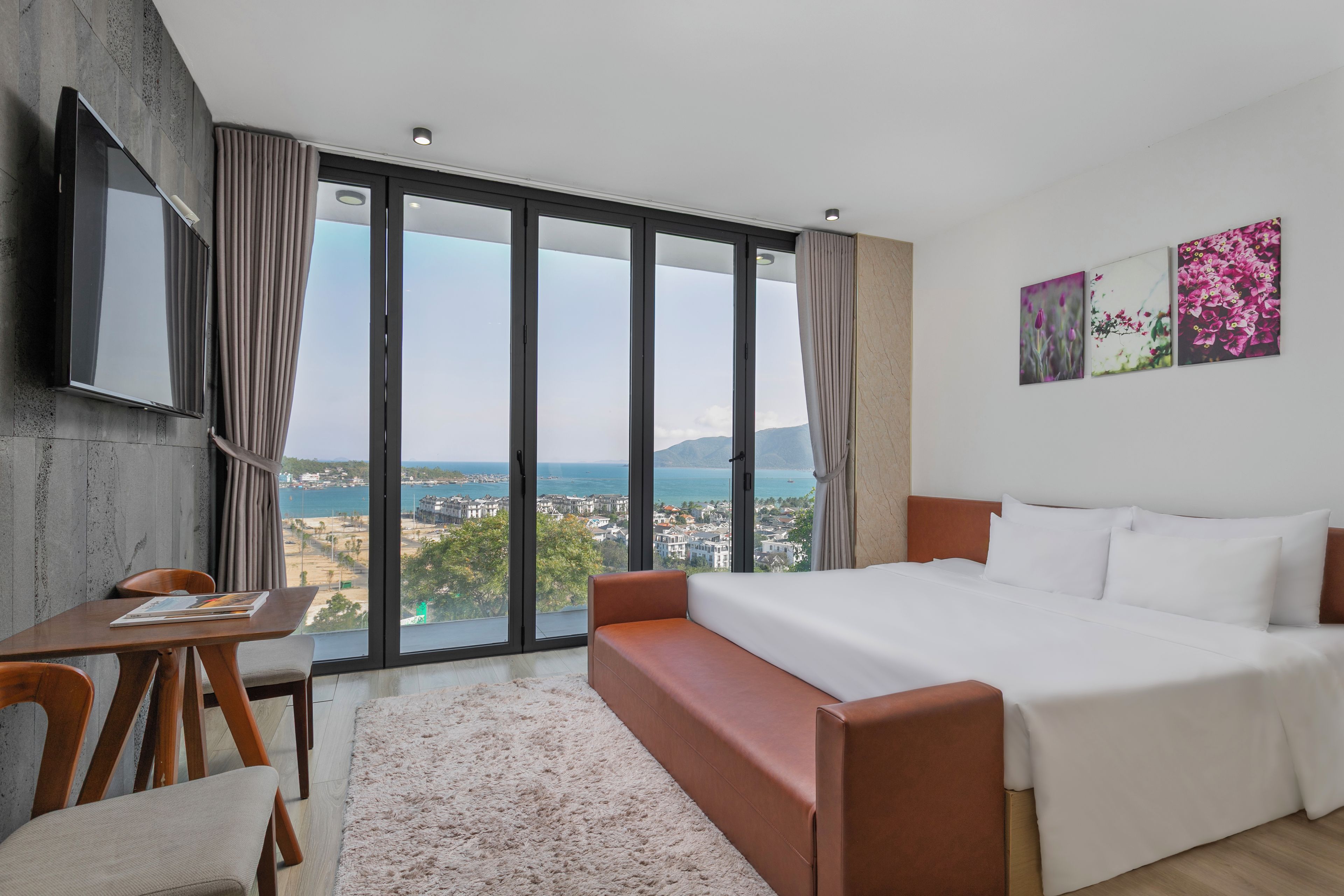 Habitación de hotel con cama, escritorio, silla, televisión y un amplio ventanal con vistas al mar y edificios.
