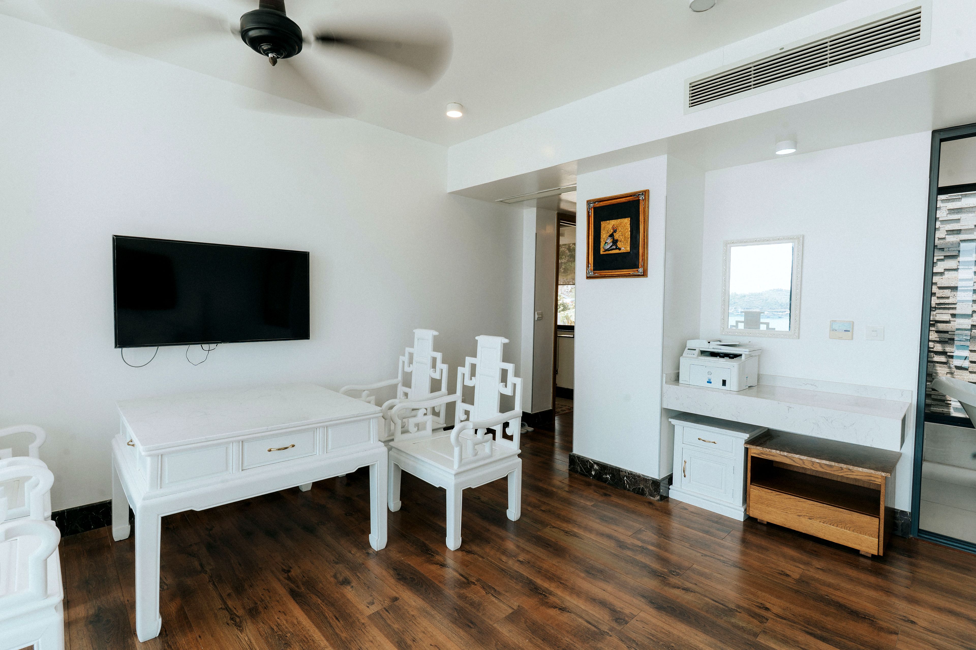 A room with a flat-screen TV mounted on the wall, a ceiling fan, a desk with a chair, and a printer on a counter.