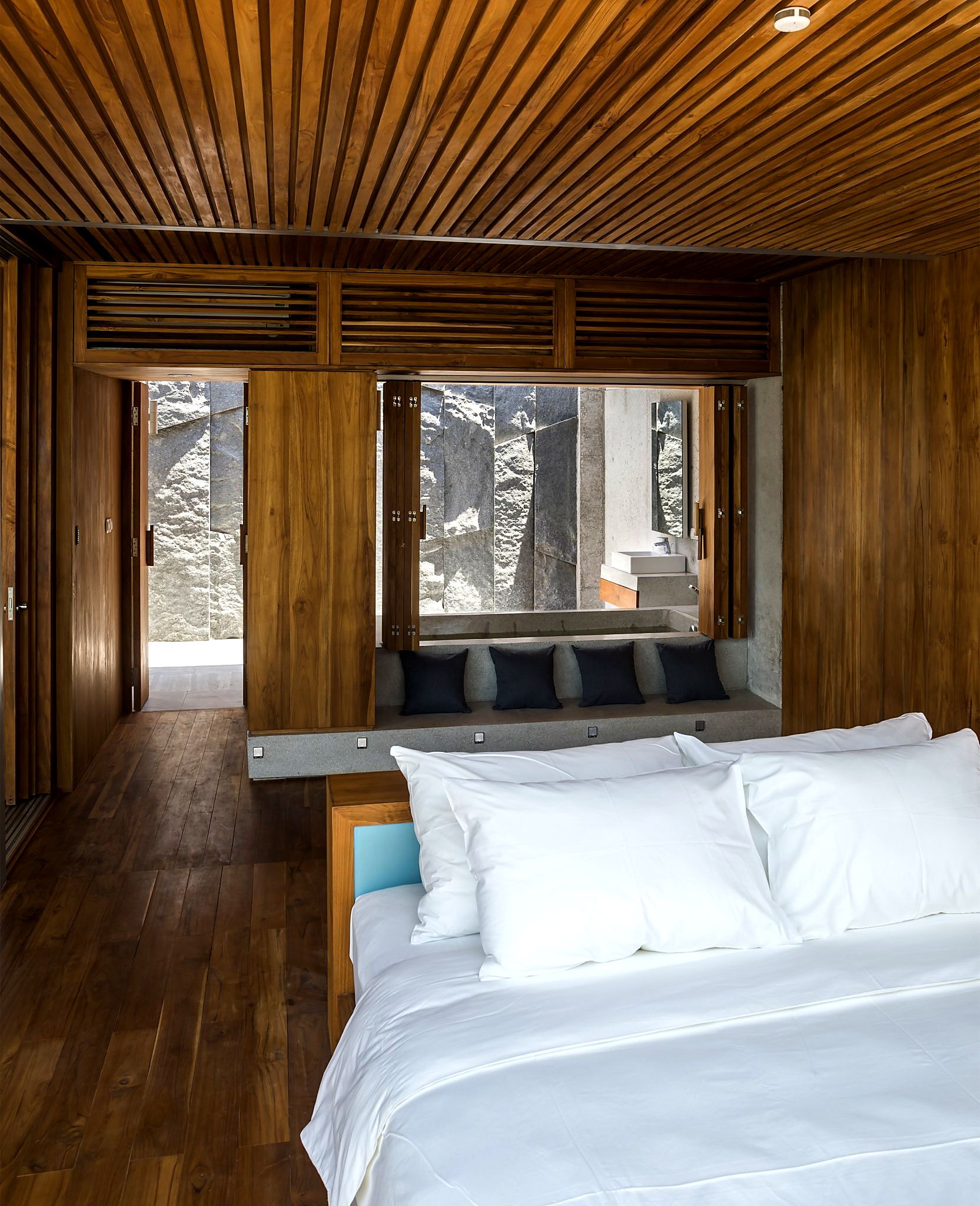 Un dormitorio moderno con techo de madera, una cama grande con ropa blanca, un banco con cojines y vistas a un paisaje nevado a través de amplios ventanales.
