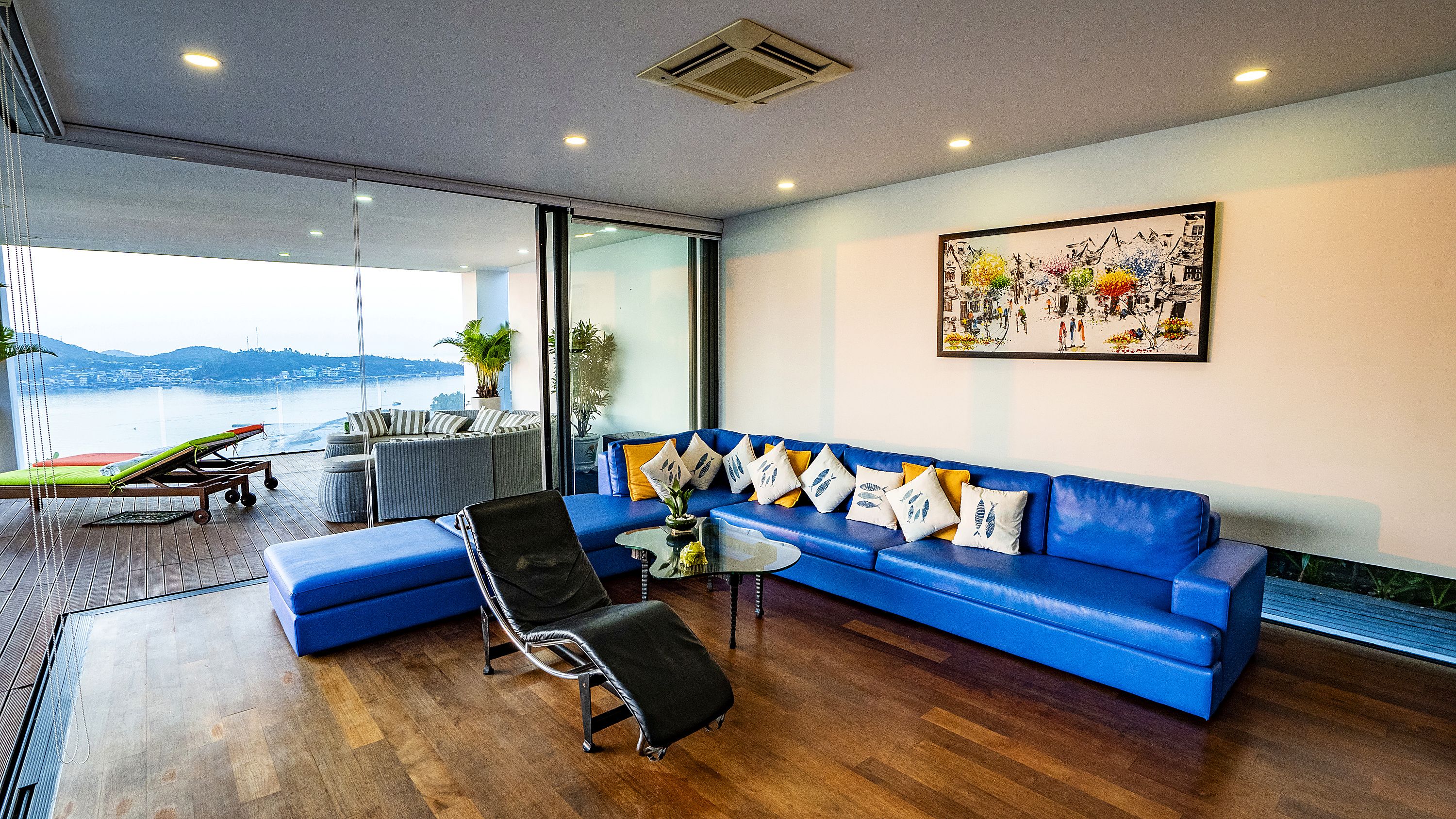 Ein modernes Wohnzimmer mit einem blauen Sofa, einem Lounge-Sessel und Blick auf ein Gewässer durch große Fenster.