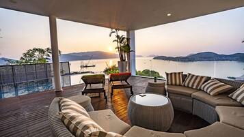 A balcony with a sofa, chairs, and a table, overlooking a body of water and distant mountains.