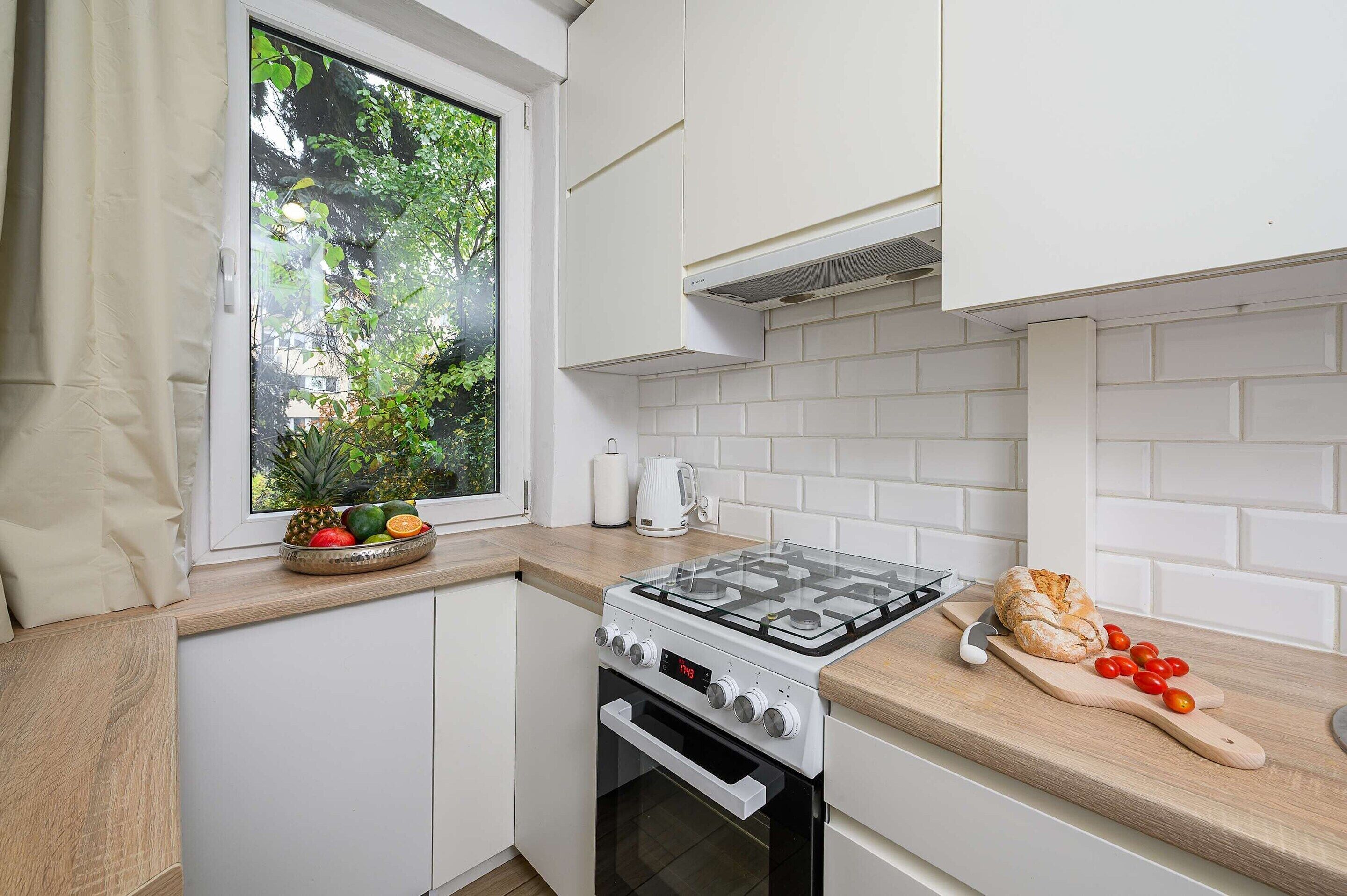 Eine kleine Küche mit weißen Schränken, einem Gasherd, einem Fenster mit Blick auf Grünflächen und einer Arbeitsplatte mit Brot und Tomaten.