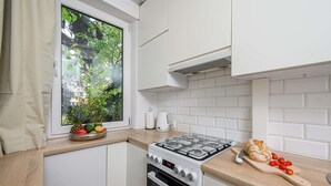 Uma cozinha compacta com armários brancos, fogão a gás, janela com vista para o verde e bancada com um pão e tomates.