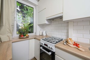 Eine kleine Küche mit weißen Schränken, einem Gasherd, einem Fenster mit Blick auf Grünflächen und einer Arbeitsplatte mit Brot und Tomaten.