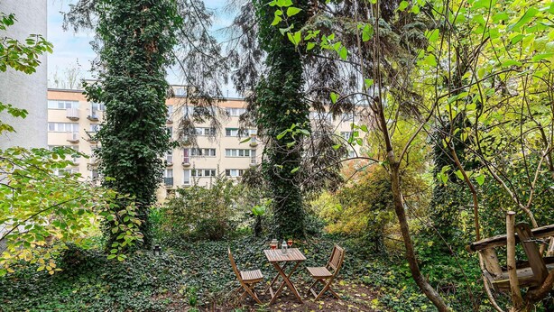 Um jardim com mesa e cadeiras de madeira, rodeado por folhagem densa e um edifício ao fundo.