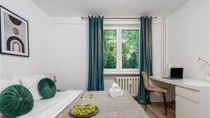 Quarto de hotel com cama, escrivaninha, cadeira, radiador e janela com cortinas.