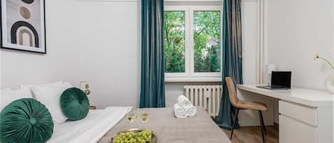 A hotel room with a bed, a desk, a chair, a radiator, and a window with curtains.