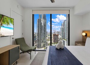 A modern hotel room with a large window, a bed, a chair, a desk, and a television displaying a beach scene. - Skyline Views Condo | Pool | Gym | Near Kaseya (Miami)