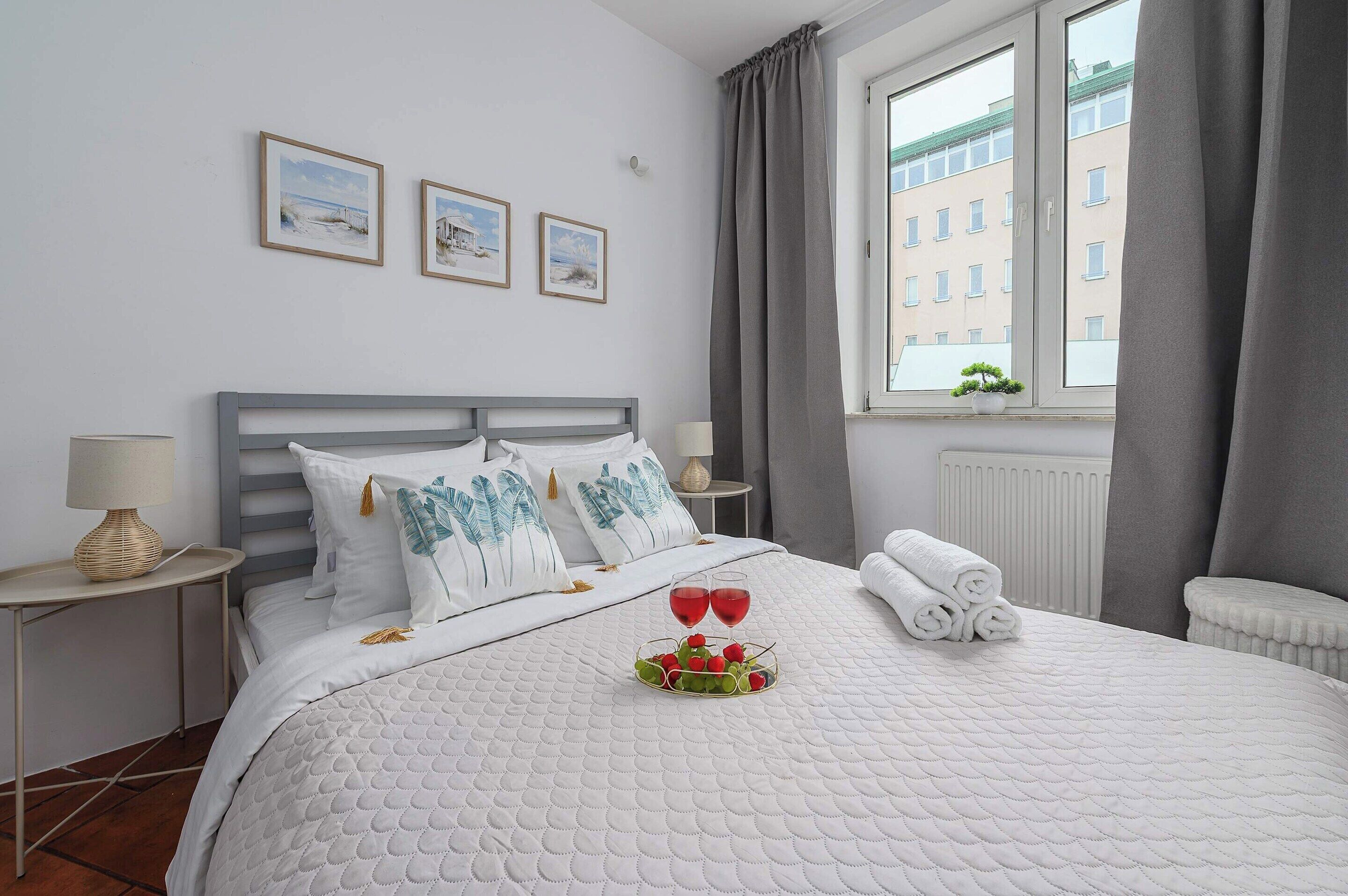 Una cama bien tendida con colcha blanca, almohadas con estampados tropicales y una pequeña composición de frutas sobre la cama.