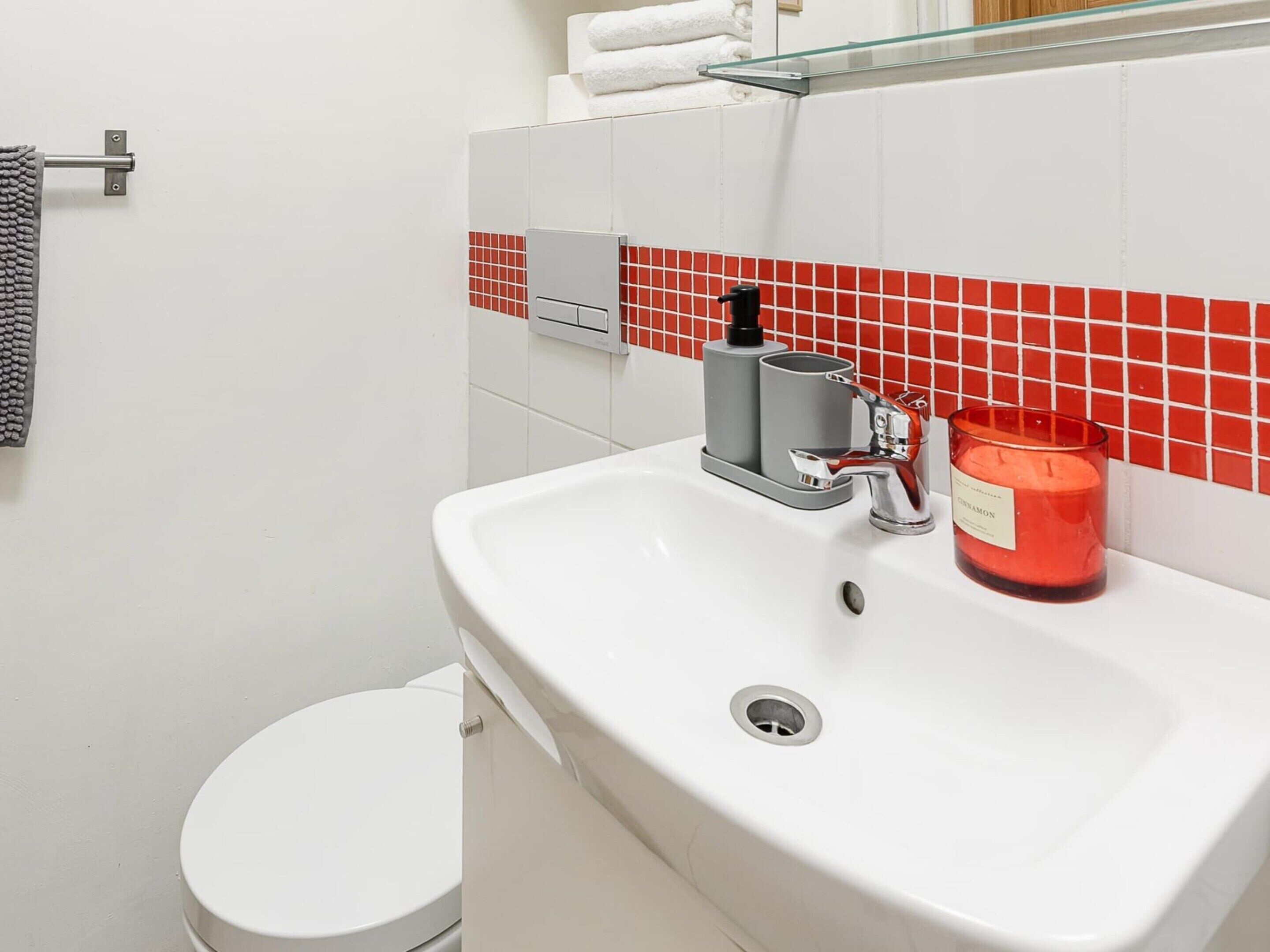 Un baño con un lavamanos blanco, un salpicadero de azulejos rojos, un estante de vidrio, un inodoro y un perchero para toallas.
