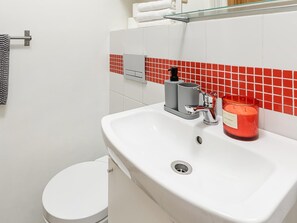 Un baño con un lavamanos blanco, un salpicadero de azulejos rojos, un estante de vidrio, un inodoro y un perchero para toallas.